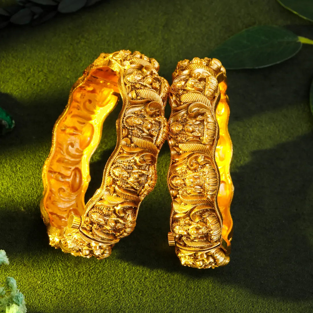 Pair of gold bangles with intricate designs on a green leafy background
